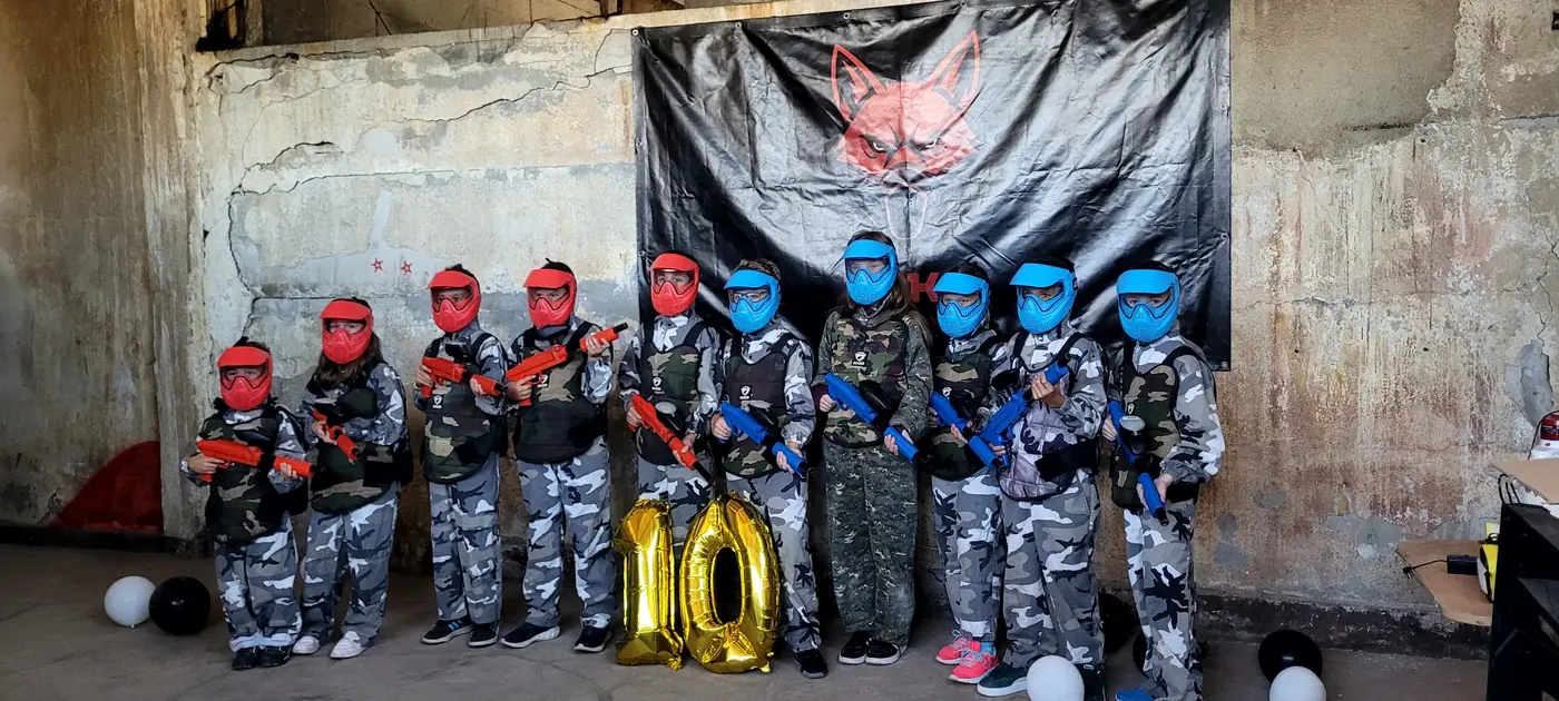 Grup de copii în costume albe de camuflaj, cu măști roșii și albastre, la o aniversare, în fața bannerului FOKS