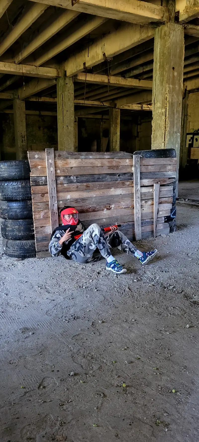 Copil în costum de camuflaj alb, cu mască roșie, așezat lângă o baricadă din lemn și cauciucuri