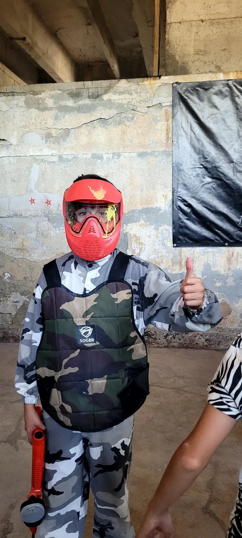 Copil cu mască roșie și vestă, cu degetul mare ridicat, într-o hală, cu markerul în mână
