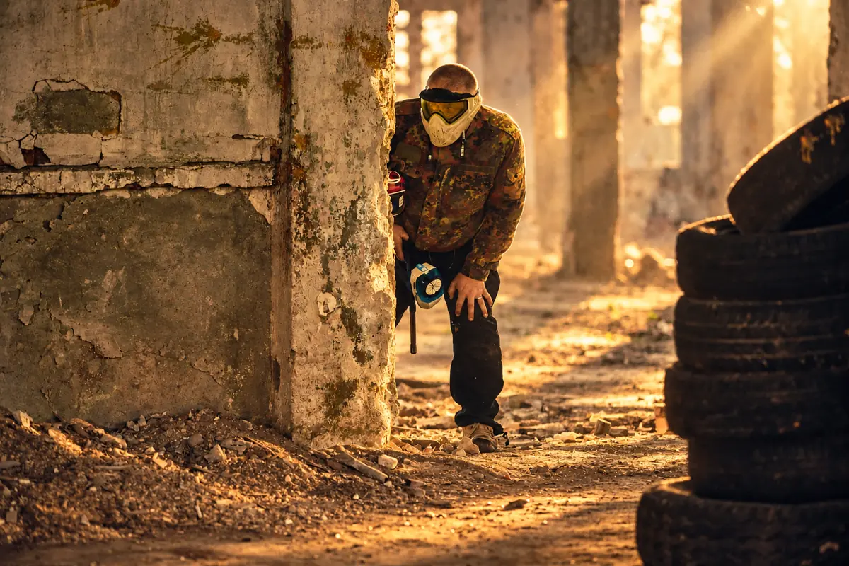 Jucător în echipament de camuflaj, aplecat după un stâlp de beton, în lumina apusului, lângă o stivă de cauciucuri